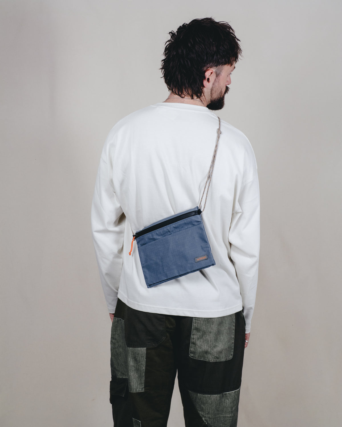 A man viewed from behind wearing a white long-sleeve shirt and dark patchwork cargo pants, with a blue crossbody pouch bag worn across his back. The bag has a thin silver-toned strap and a small brown leather label.