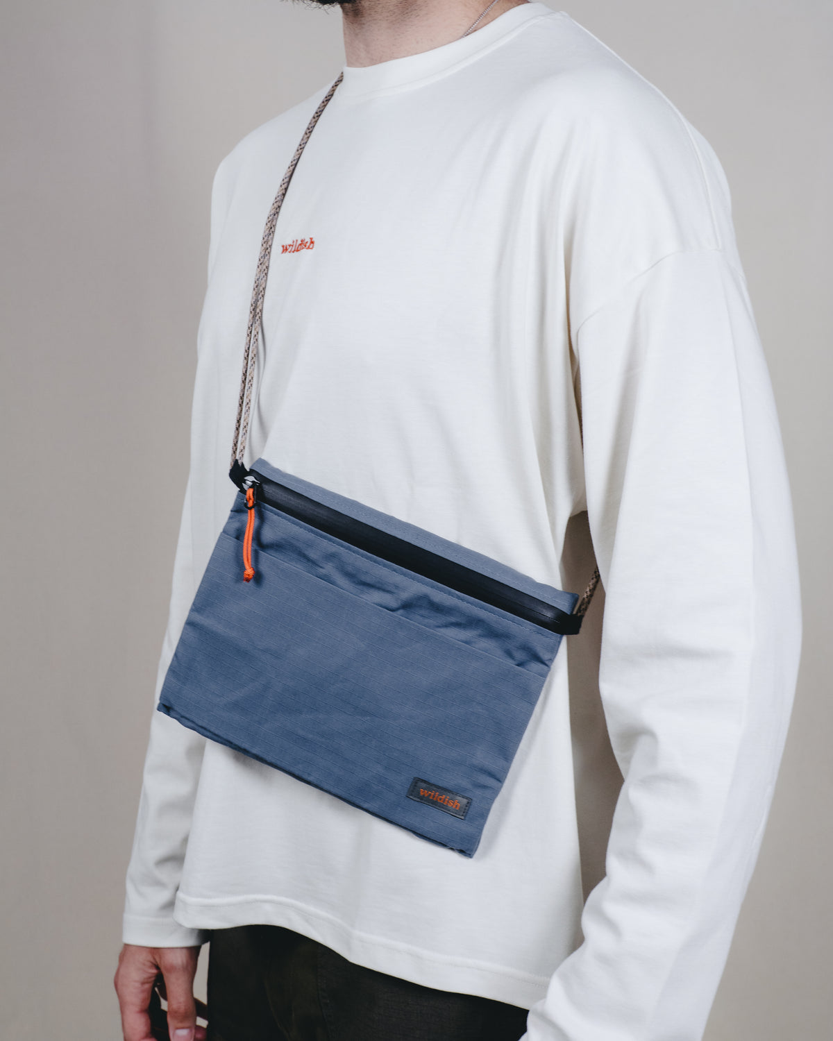 A person wearing a white long-sleeve shirt carries a blue rectangular crossbody pouch bag with a black zipper, orange zipper pull, and a brown woven strap against a plain light gray background.