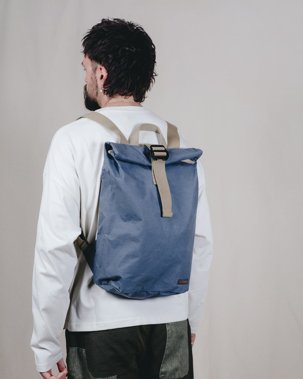 A man viewed from behind wearing a blue roll-top backpack with beige straps and trim, styled with a white long-sleeve shirt and dark patchwork pants against a plain beige studio backdrop.