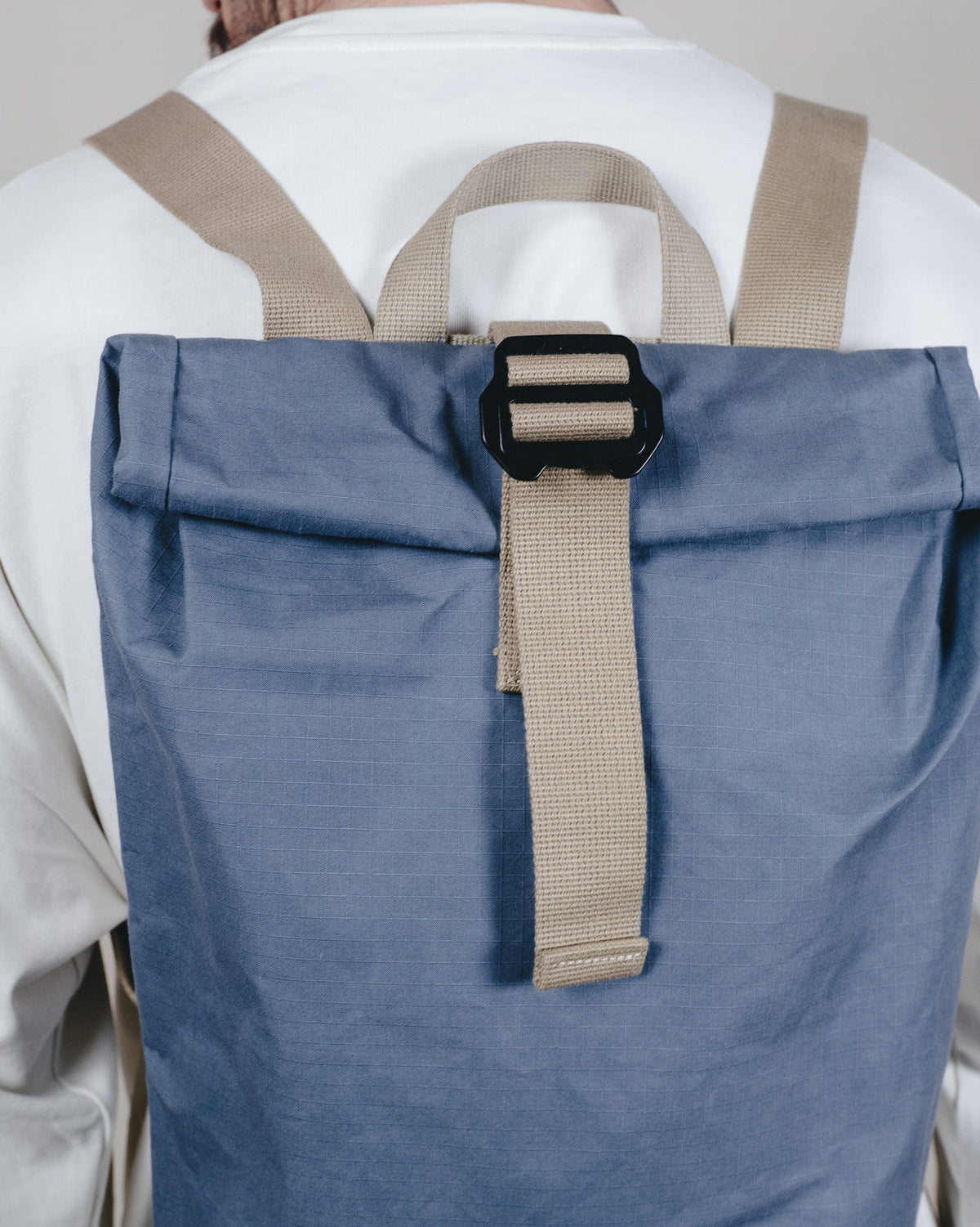 A close-up back view of a person wearing a roll-top backpack in dusty blue with beige straps and a black buckle.