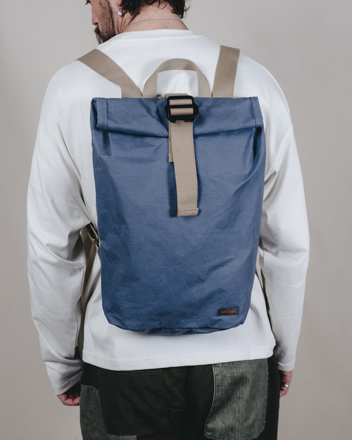 A man viewed from behind wearing a blue roll-top  backpack with beige straps and a black buckle, over a white long-sleeve shirt and dark pants.