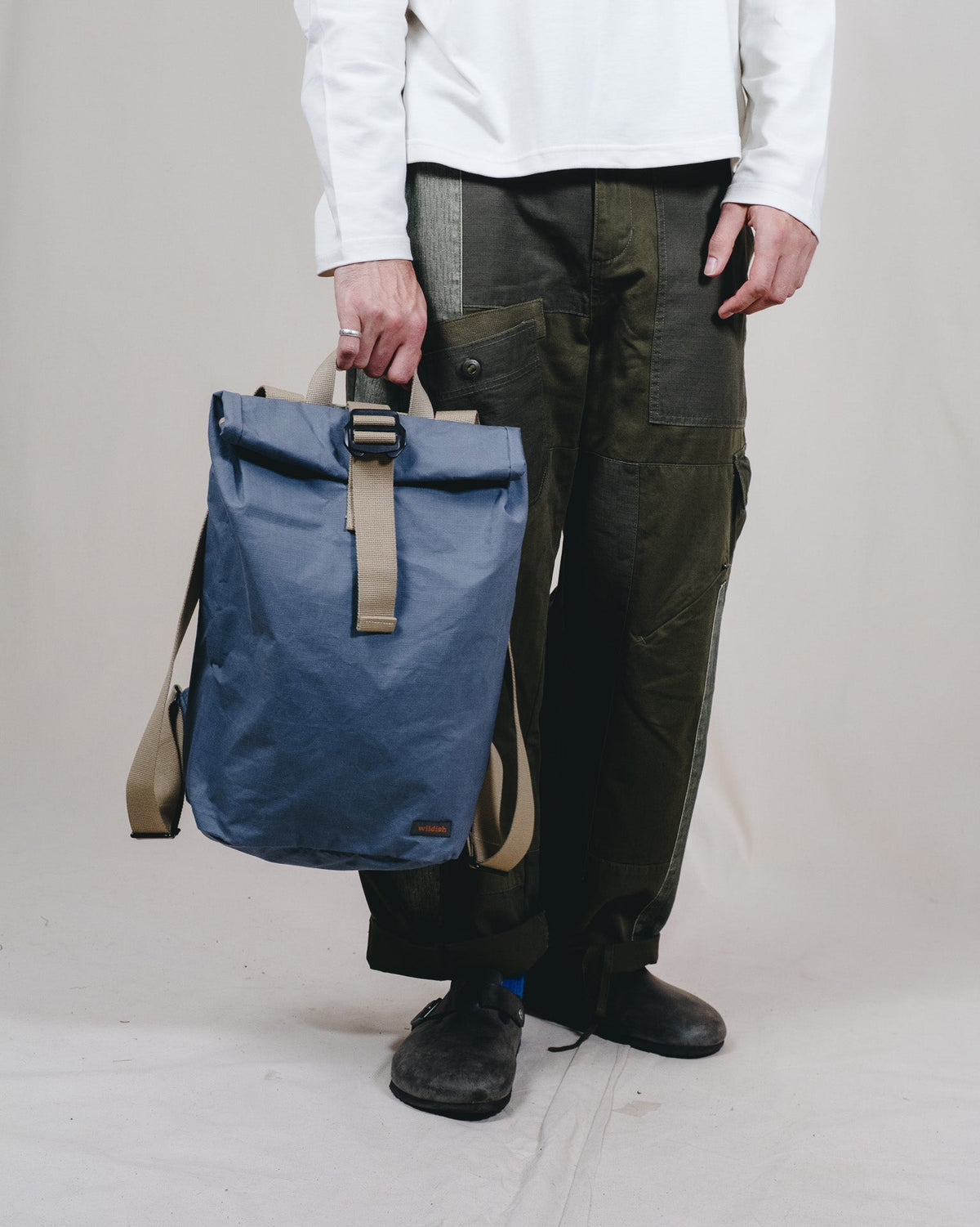 A person wearing a white long-sleeve shirt, dark olive cargo pants, and black shoes holds a blue roll-top backpack with beige straps against a plain light gray studio background.
