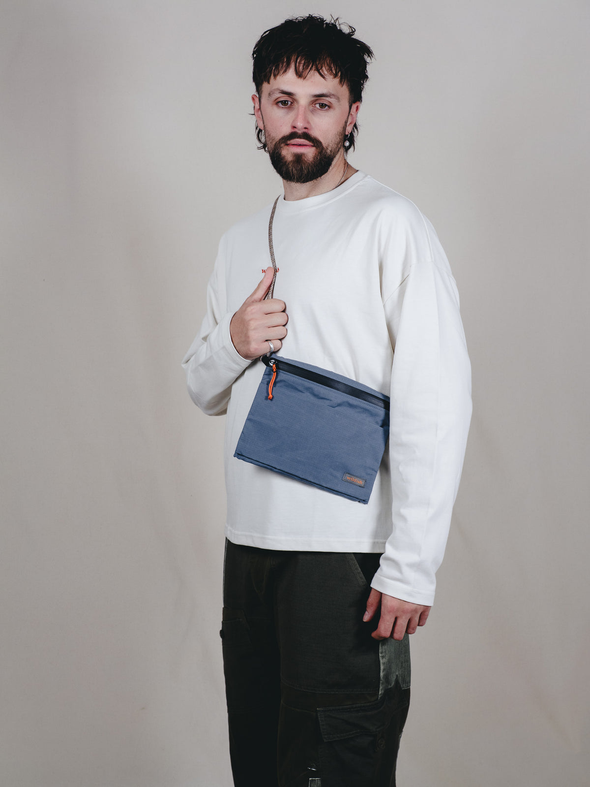 A man wearing a white long-sleeve shirt and black cargo pants models a blue crossbody pouch bag with a thin strap and zipper closure against a plain beige studio background.