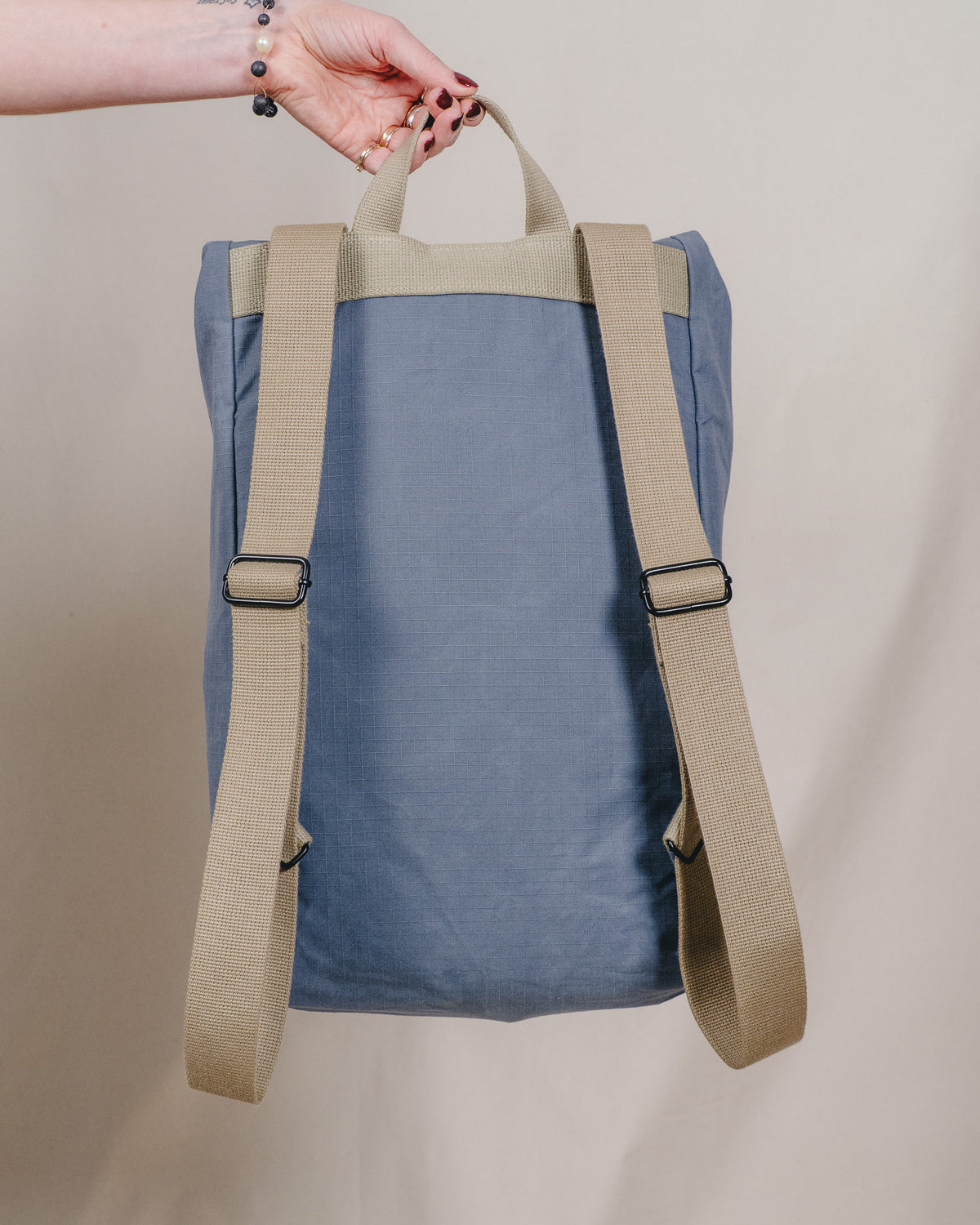 A blue fabric backpack with beige webbing straps and top handle, held by a person's hand against a plain beige background.