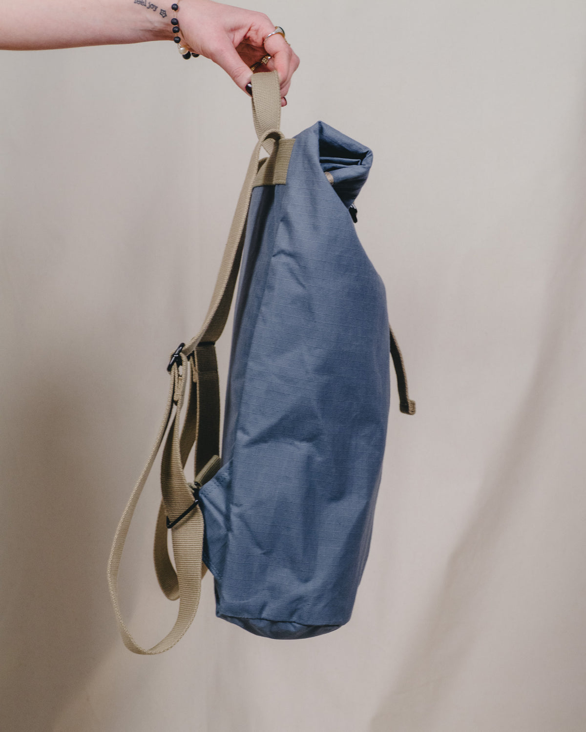 A person's hand holding a blue roll-top backpack with beige straps against a plain beige background.
