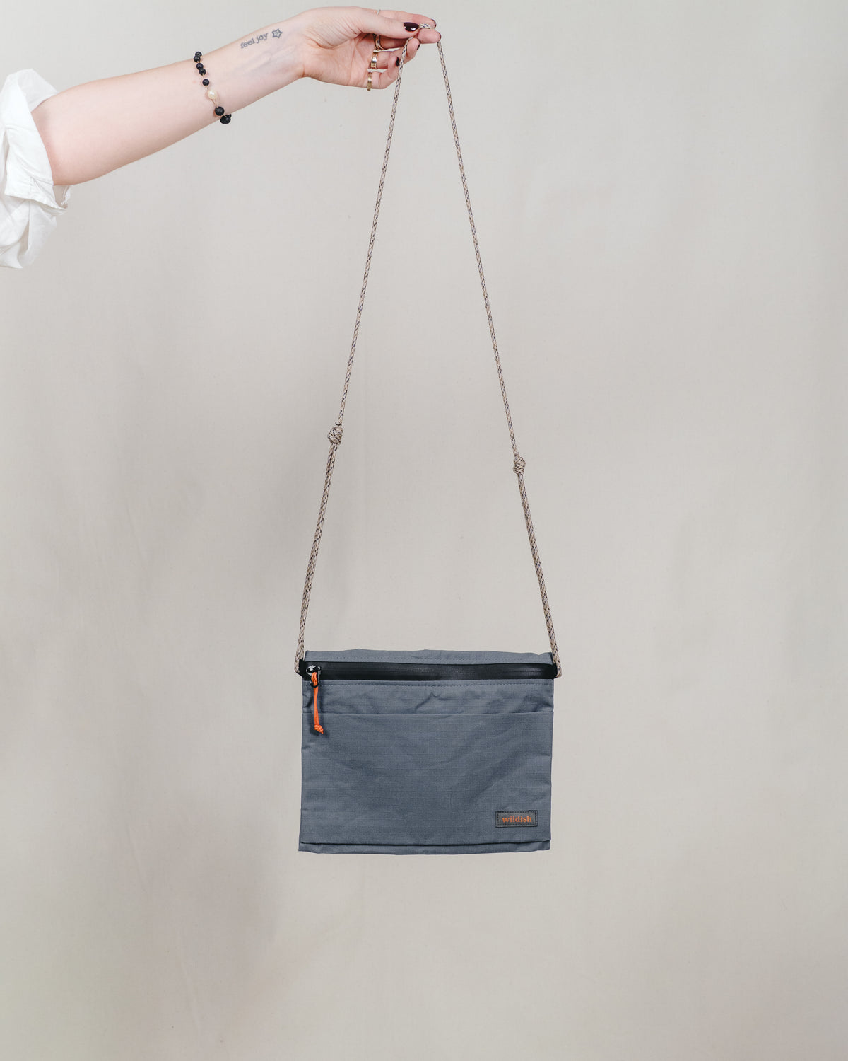 A slate blue rectangular crossbody bag with a black top trim, held by a person's hand against a plain beige background.